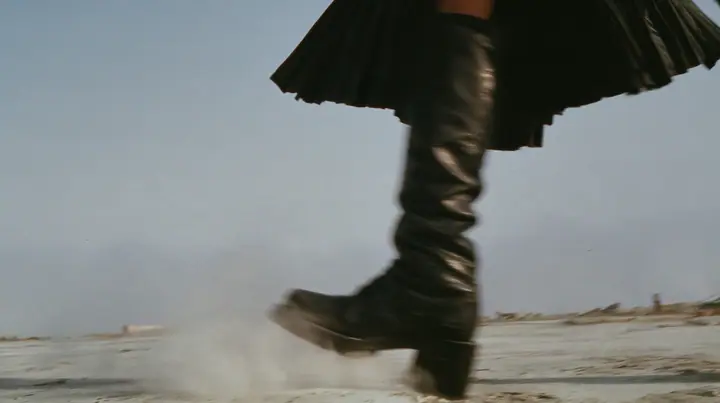 low close-up of a black knee boot stamping down on pale concrete, a puff of dust, a pleated black skirt hem at the top of the frame, no face, pale dawn grey-white, soft even light, a sea of fog below, one small…