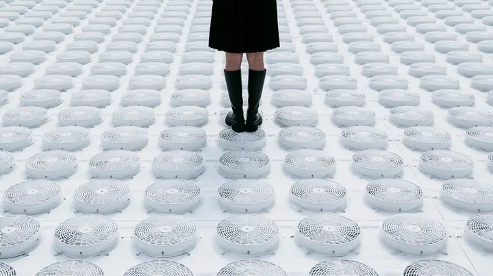 straight-down aerial of a vast datacenter roof with hundreds of identical round cooling fans in a grid like a city, white haze, the black pleated skirt hem and two black knee boots at the top edge of the frame,…