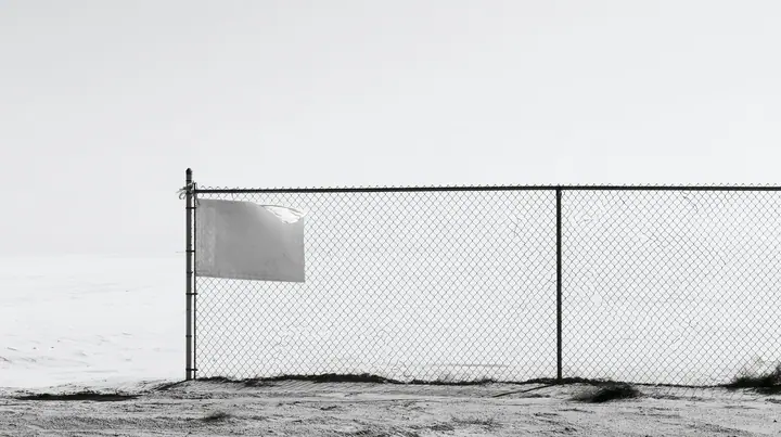 wide shot of a chain-link construction fence at the edge of a vast empty desert site, one blank laminated notice cable-tied to it and flapping, high-key white sky, no people, high-key white paper light, almost no…