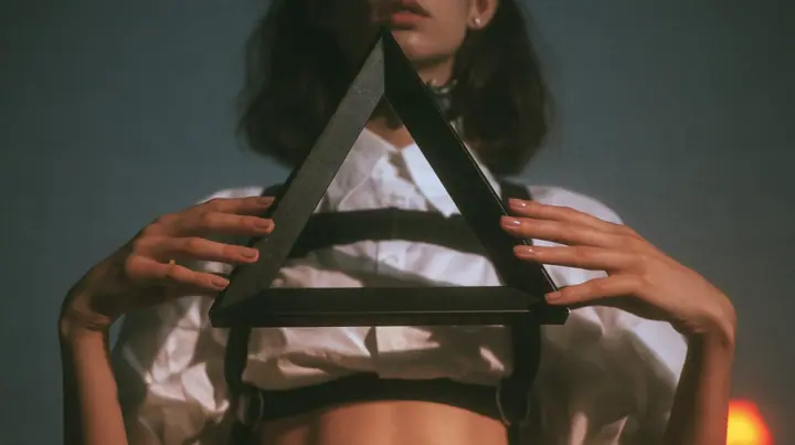 close-up of a young Caucasian American woman's hands holding an impossible triangle object made of matte black metal at chest height, a white cropped shirt and harness straps behind, no face, the right third empty…