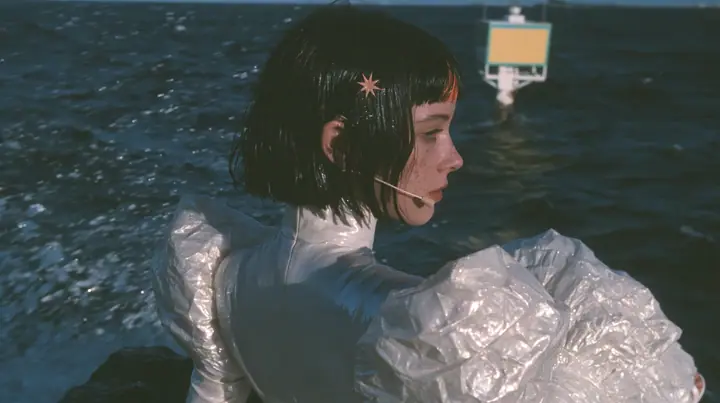 over-the-shoulder shot from behind a Caucasian American woman in her mid-twenties with pale skin and light freckles, a glossy black blunt jaw-length bob with heavy straight bangs and one clay-orange streak through…