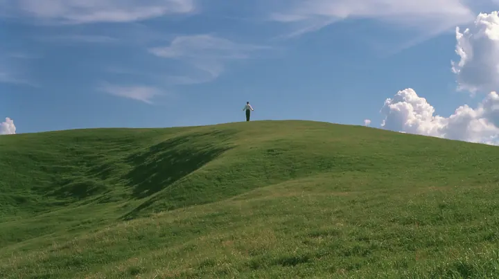 Y2K high fashion editorial photograph, 2001: a vast wide shot of a smooth rolling bright-green grassy hill with soft curves, the grass short and velvety with a tiny lone figure standing on the crest seen from behind,…
