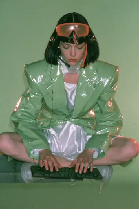 Y2K high fashion editorial, a 2001 couture magazine story: full-length shot of a young Caucasian American woman with pale skin and a glossy black blunt bob with heavy straight bangs and one clay-orange streak, a thin…